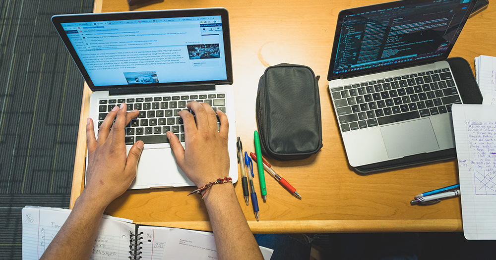 Two laptops are open on a desk; one set of hands types on one laptop. There are pens and papers around.