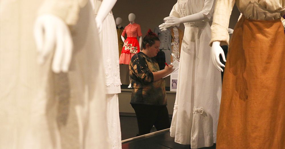 An english student wanders through a room of women's suffrage dresses in a museum while doing a writing exercise