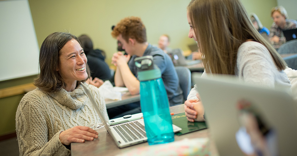 A communications professor chats with a student in an active classroom
