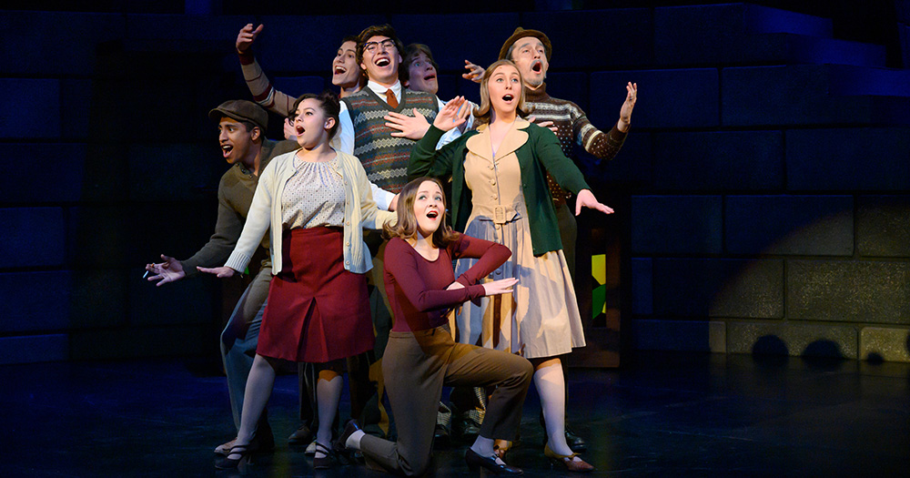 Theatre students in mid-performance mode, singing and posing on a lit stage.