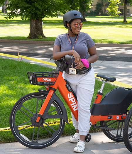 A student wearing a helmet and sunglass smiles while leaning on a Spin rentable E-bike on the green, sunny oval at Colorado State.