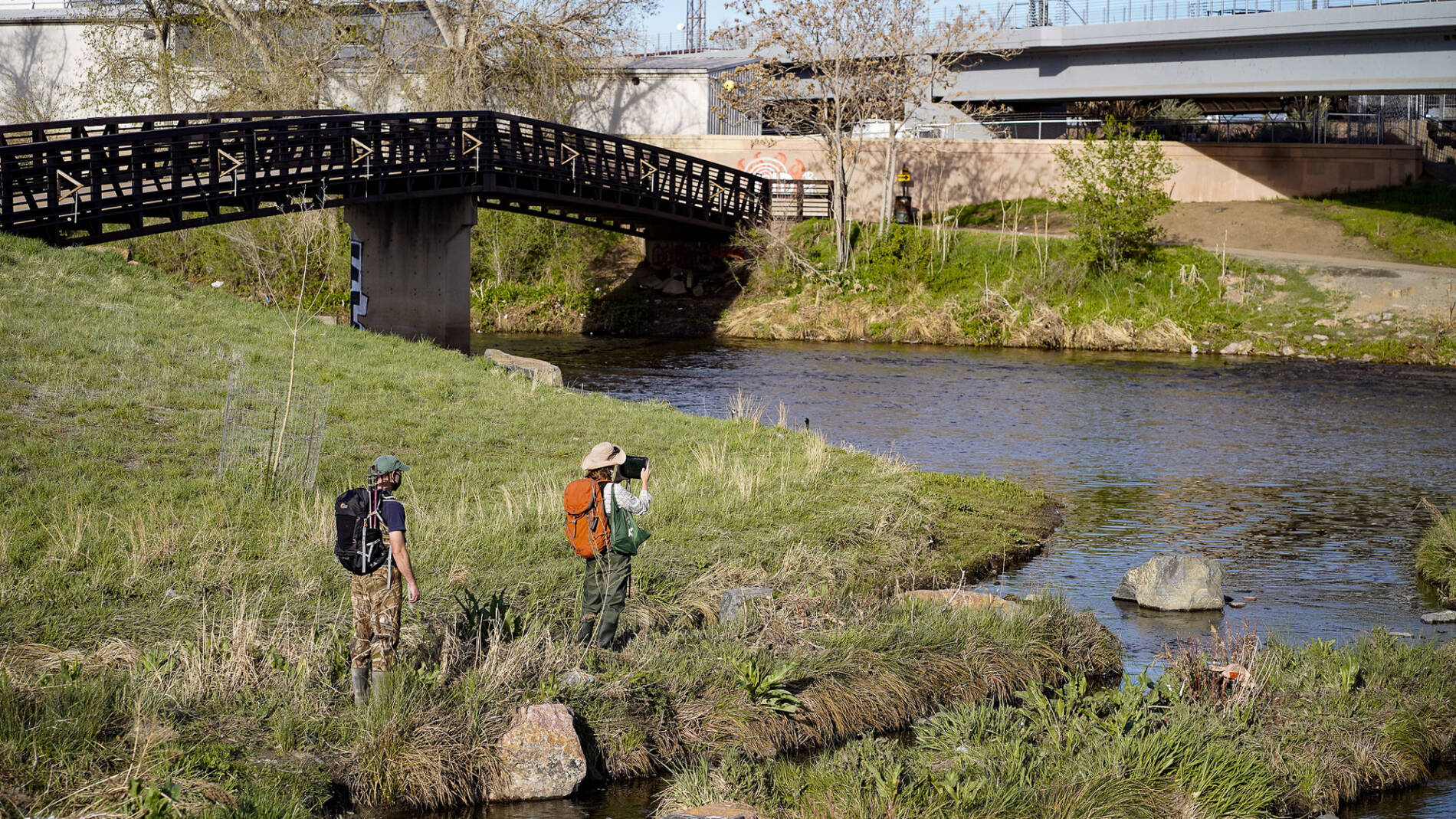Two students with hiking packs and a tablet studying a river near a bridge