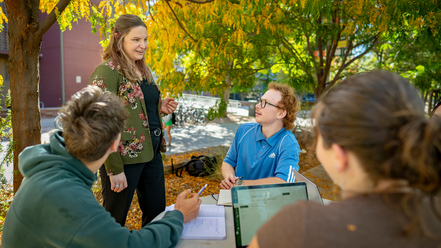 Students discuss literature outdoors in a tree-covered setting