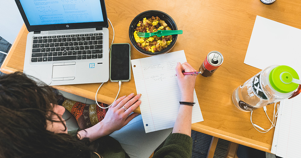 looking down at a study table with a laptop, phone, food, and drink