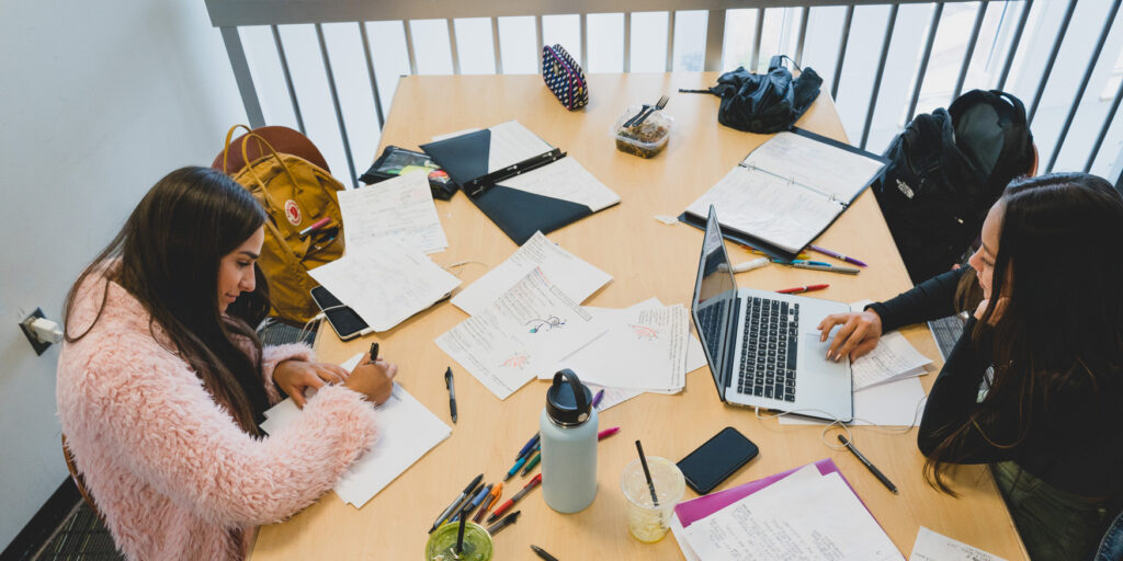 Two students with papers and food and laptops spread out on big table as they study