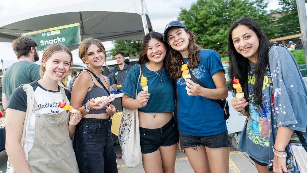 Group of students eat fruit kebabs and popcorn under a sign that says "snacks"