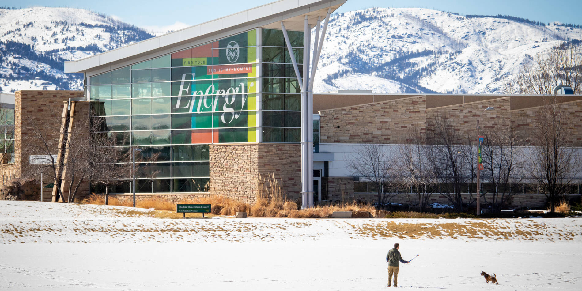 Building with huge Energy banner covering the windows sits in the foreground of the snowy foothills in the background, with snow on the ground and a man playing fetch with his dog.