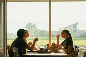 Three students sit at a table facing a window eating together