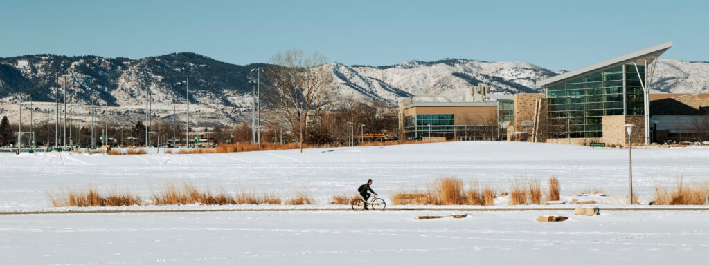 Person riding bike along sidewalk. The ground mountains in the background are covered in snow.