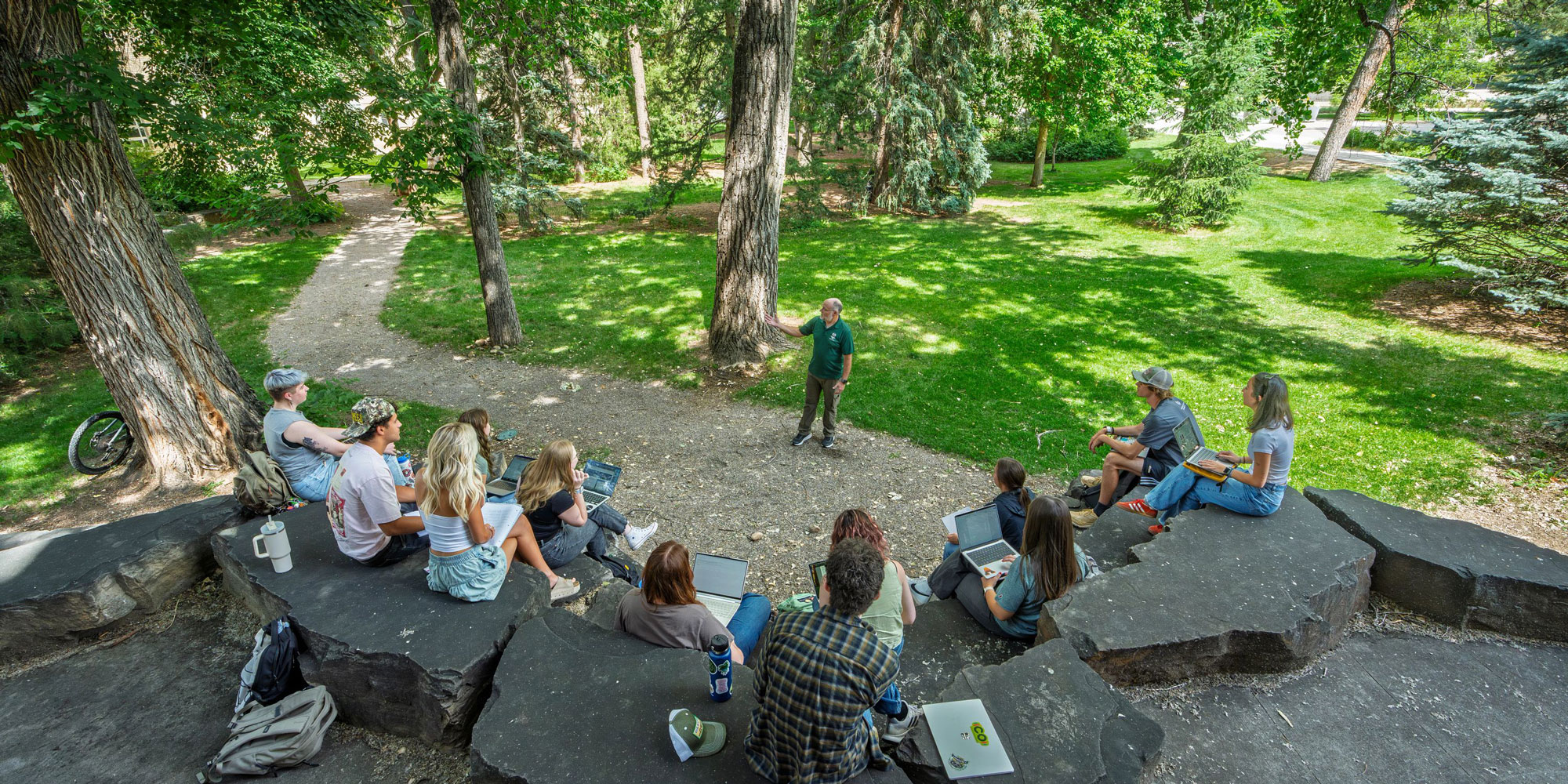 Group of students sit on rocks outside in a heavily treed area while a professor speaks to them