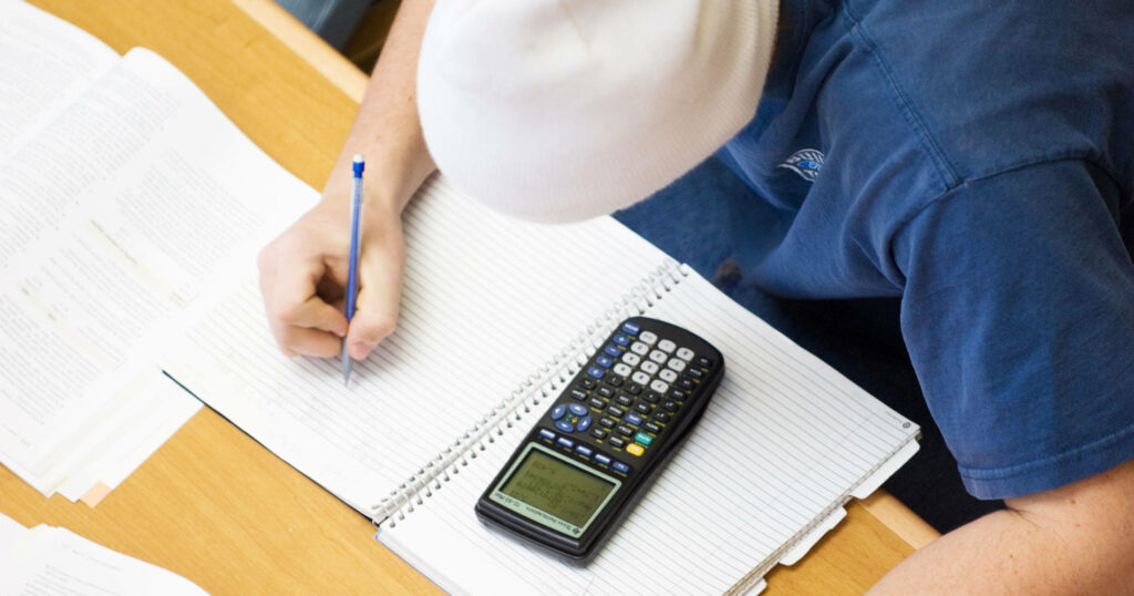 A student writes in a notebook while using a calculator.