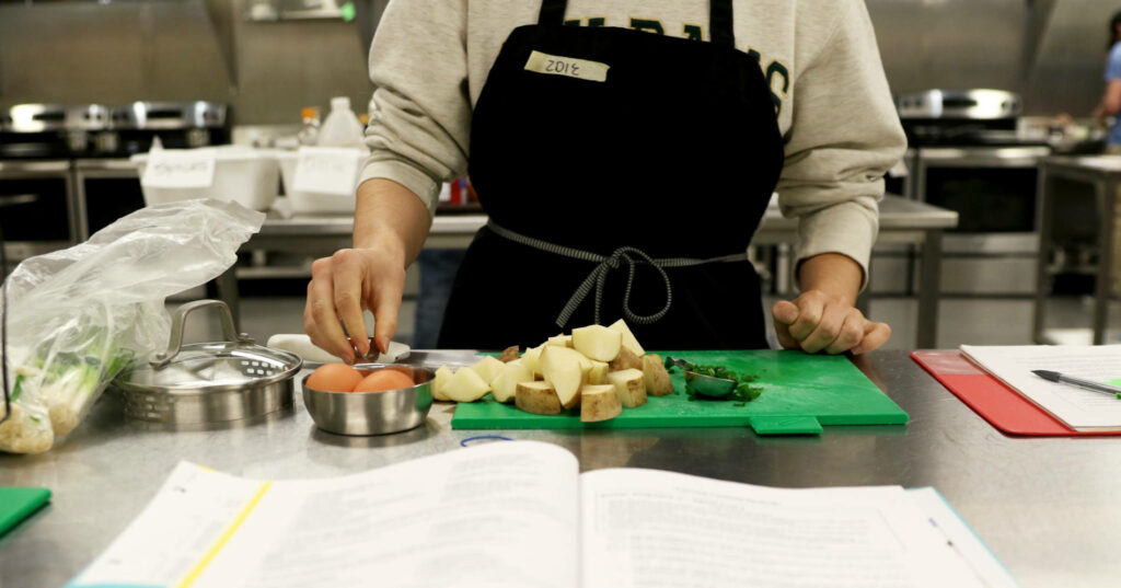 Closeup of a student cooking fresh food in a teaching kitchen. Open book nearby.