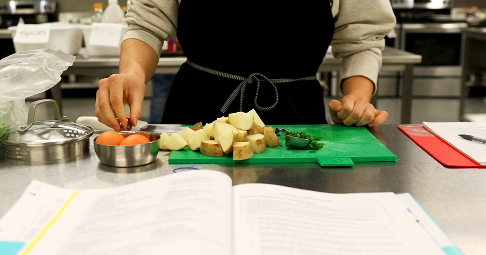 A close-up of hands chopping fresh food in a professional kitchen. Recipe books, class notes, and other ingreidents sit nearby.