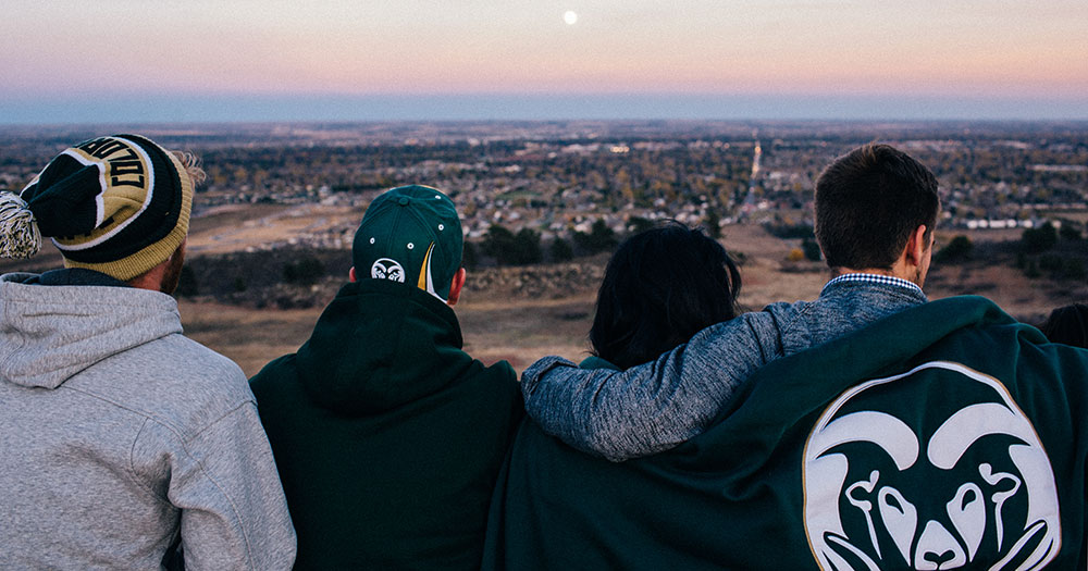 Four students wearing CSU gear sit closely on a ridgeline and enjoy a sunset and a view of Fort Collins.