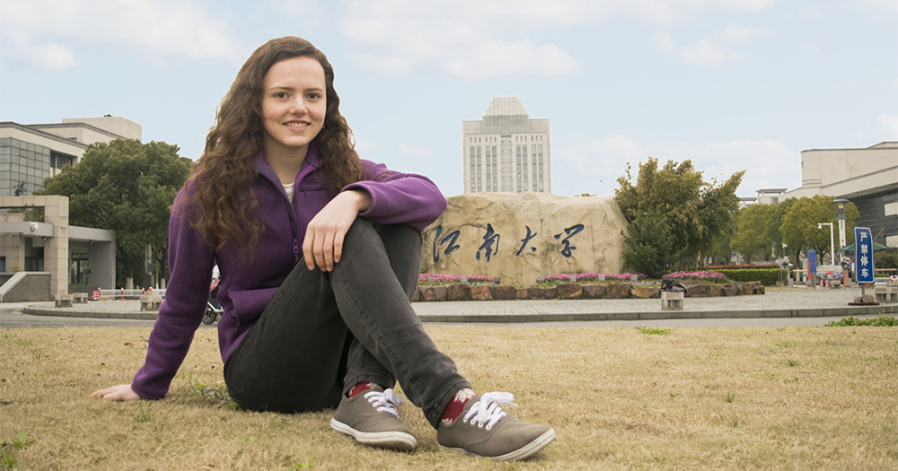 mary swing sits cross-legged on the ground and poses for a photo in front of city buildings and a large stone carved with chinese characters