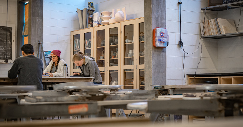 Colorado State pottery students work in the wide-open studio space with tools and shelves of pottery behind them