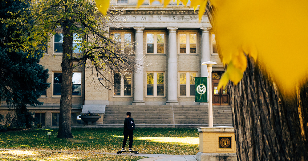 A students glides by the administration building on a skate board during a bright fall day at Colorado State.
