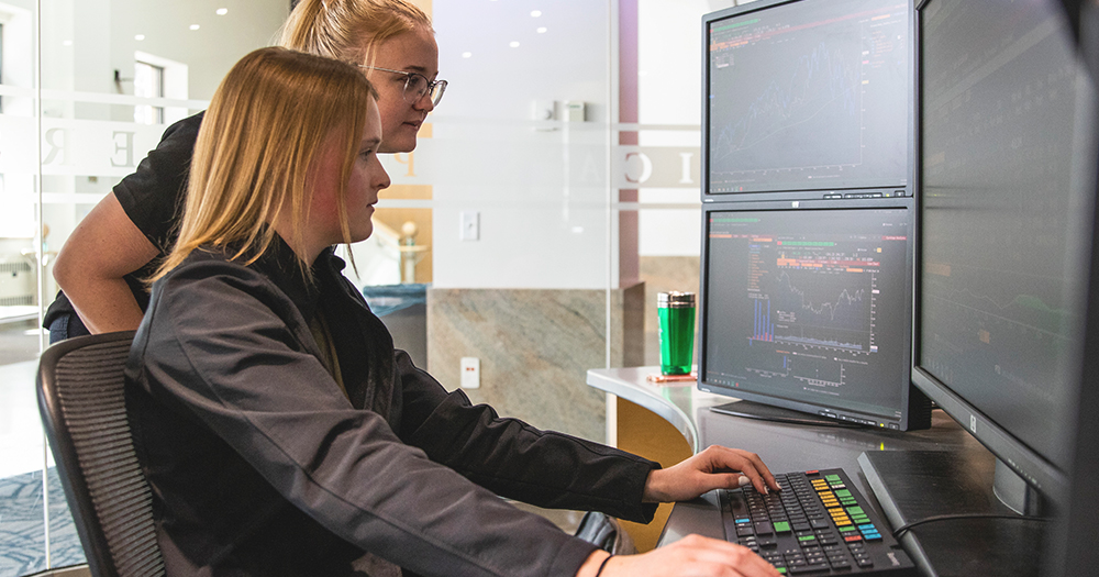 Two students work with data on big stock market screens