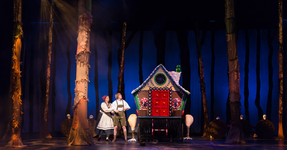 two theatre students on stage during a hansel and gretel performance. trees and a decorative witch cottage included