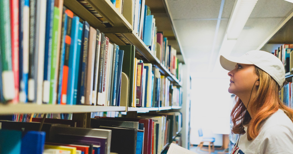 A student gazes up at a library shelf of books.