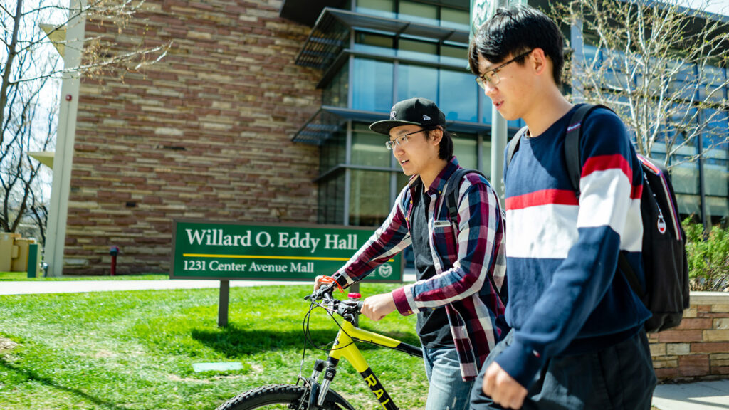 Two international students walk across campus. One is walking with his bike.