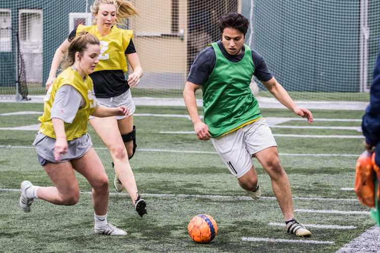 Two women and one man playing indoor soccer and wearing green and yellow pinnies