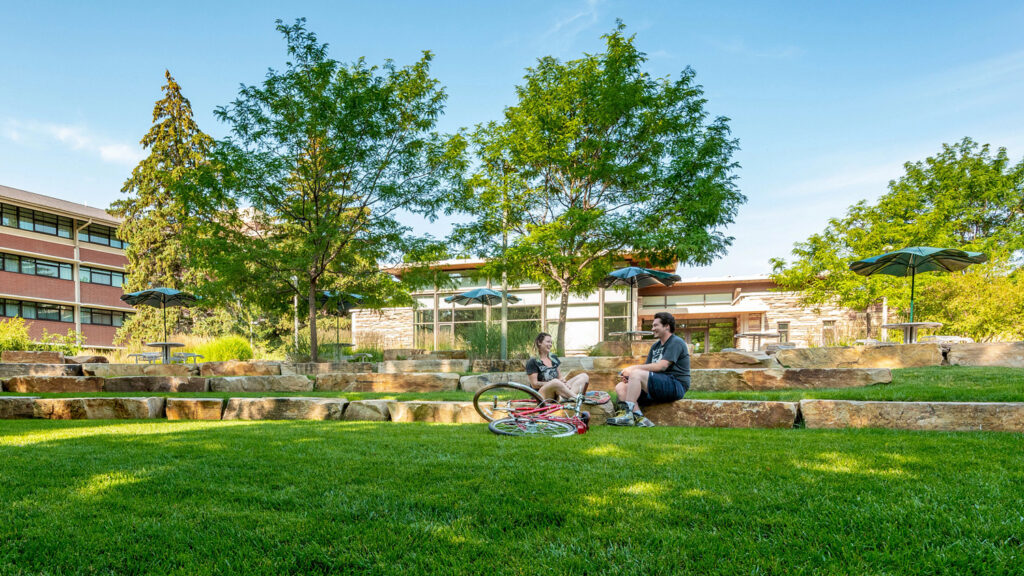 Two students talk outside with rocks, trees, and residence halls in background