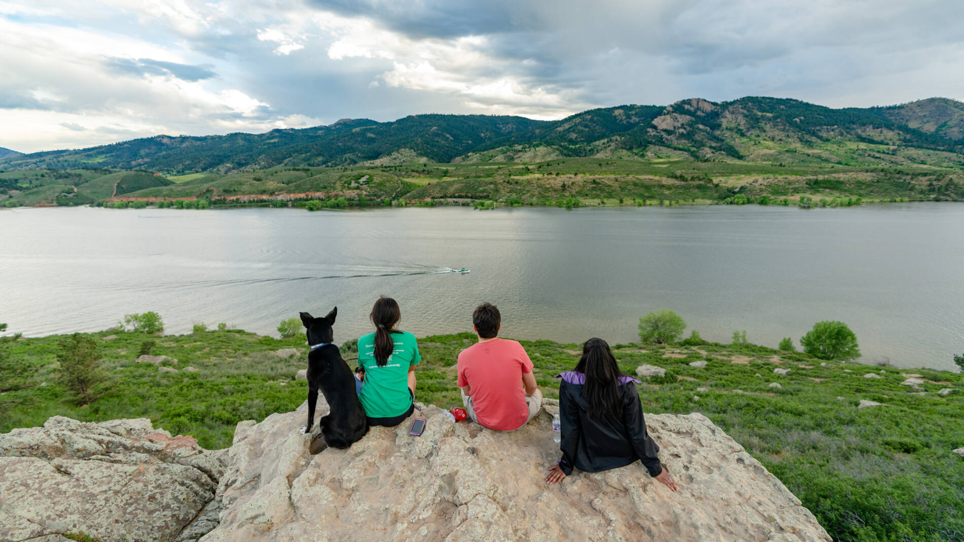 Three students and a dog sit on a rock edge looking out over a lake with a boat driving by