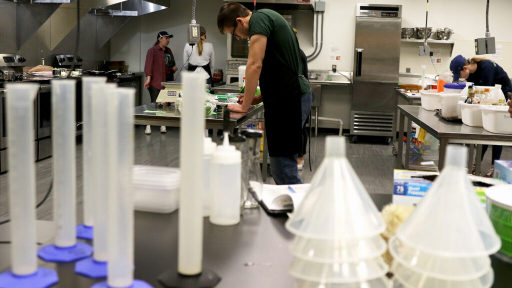 A student works on paperwork in a learning kitchen; beakers and scientific tools sit in the foreground.